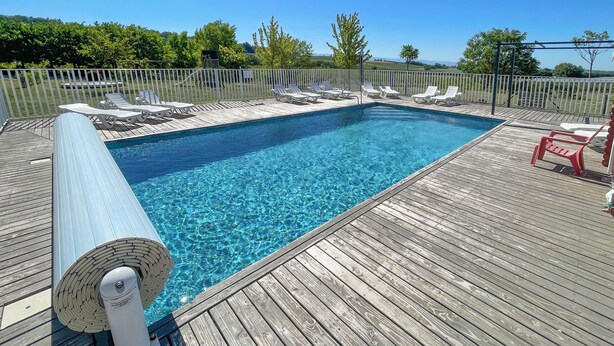 Uma piscina retangular com água azul cristalina, rodeada por um deck de madeira com espreguiçadeiras brancas e vermelhas.