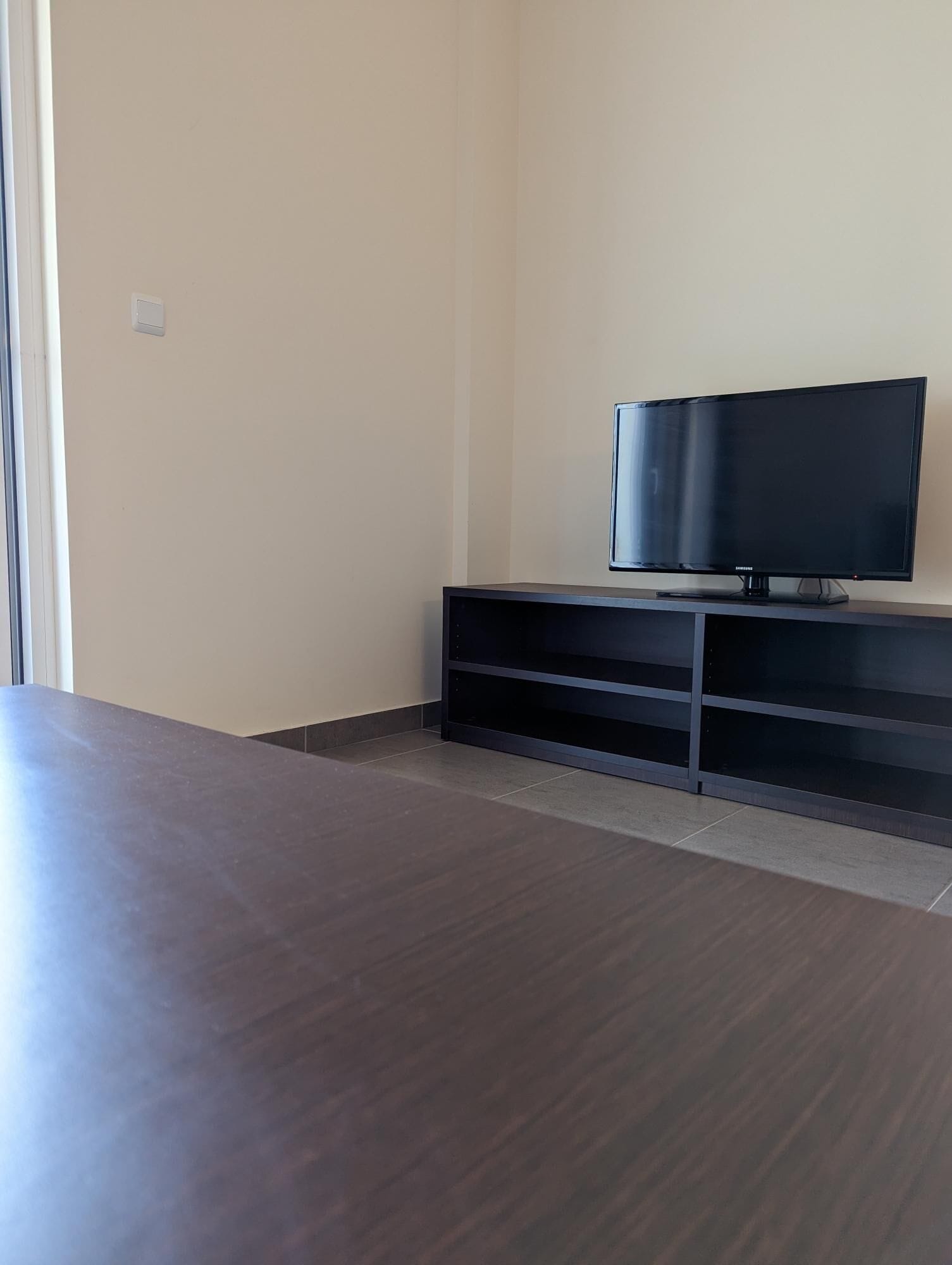 Une pièce avec un téléviseur à écran plat posé sur un meuble noir, une grande table en bois foncé et un mur de couleur claire.