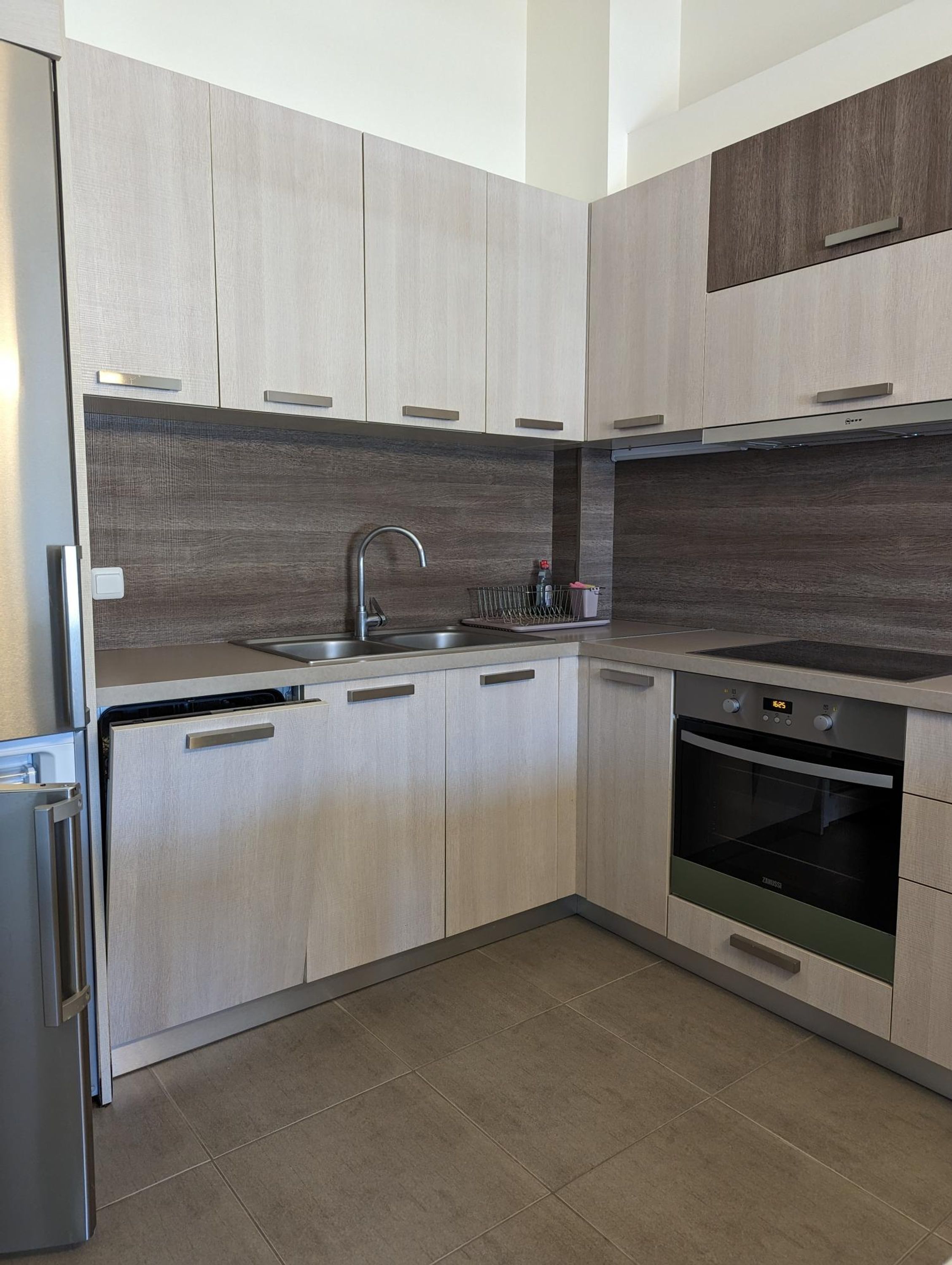 A modern kitchen with light wood cabinets, stainless steel appliances, and a tiled floor.