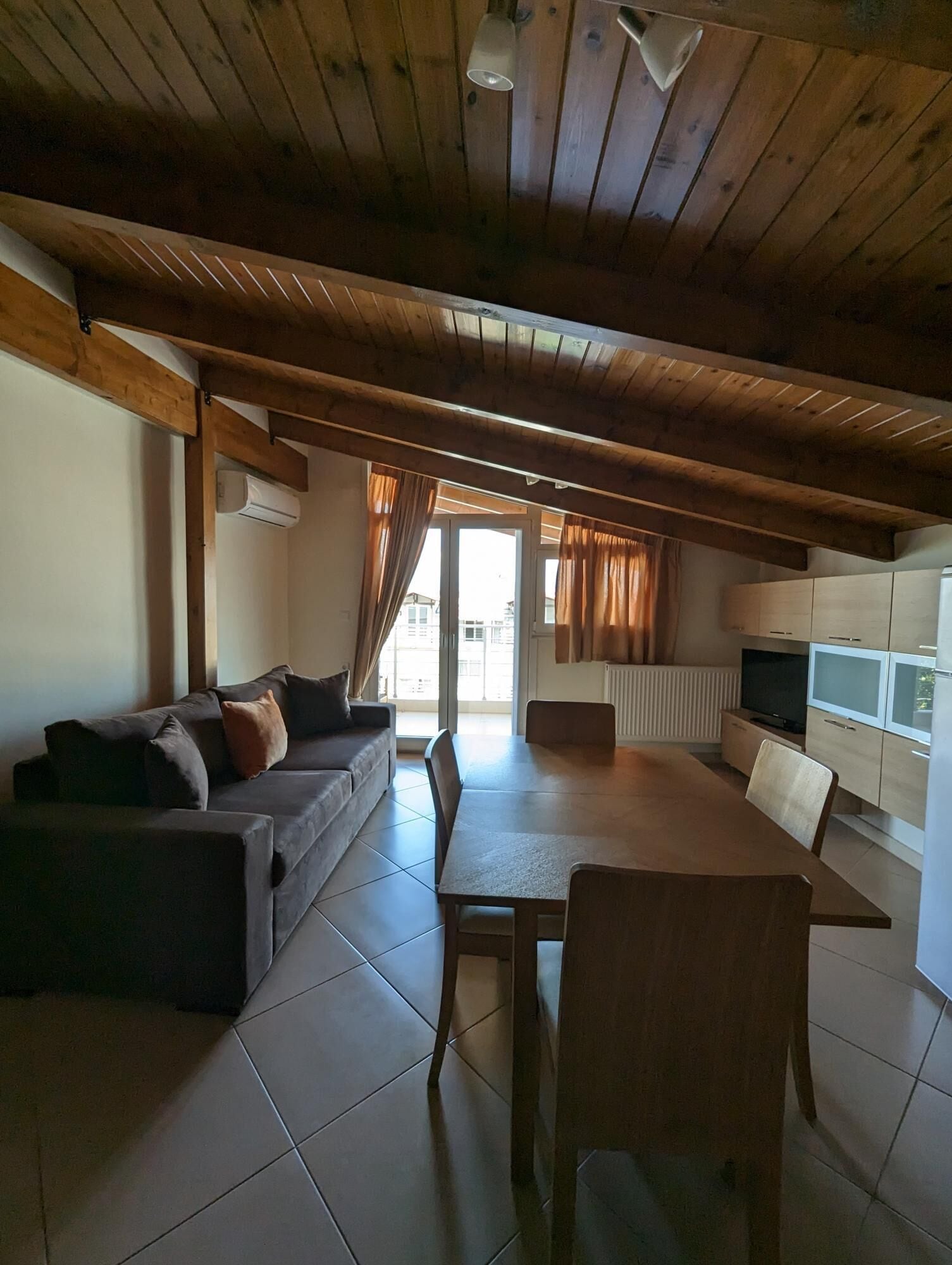 A cozy living area with a wooden ceiling, a sofa, a dining table with chairs, and a kitchen area with cabinets and a TV.