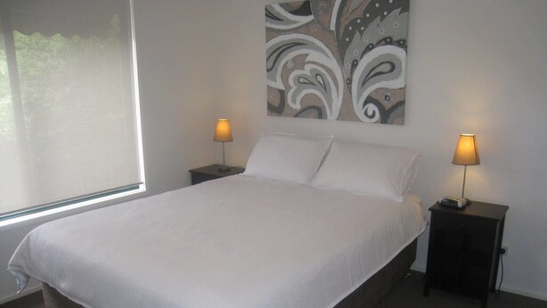 A neatly made bed with white linens, two bedside tables with lamps, and a large abstract painting on the wall. - Comfy family holiday destination (Ocean Grove)