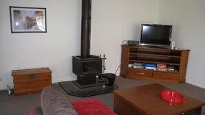 A living room with a sofa, a coffee table, a television on a wooden stand, a fireplace, and a small wooden chest. - Comfy family holiday destination (Ocean Grove)
