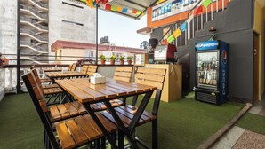 Un espace extérieur aménagé avec des tables et des chaises en bois, un réfrigérateur à boissons de la marque Pepsi et des drapeaux aux couleurs vives.