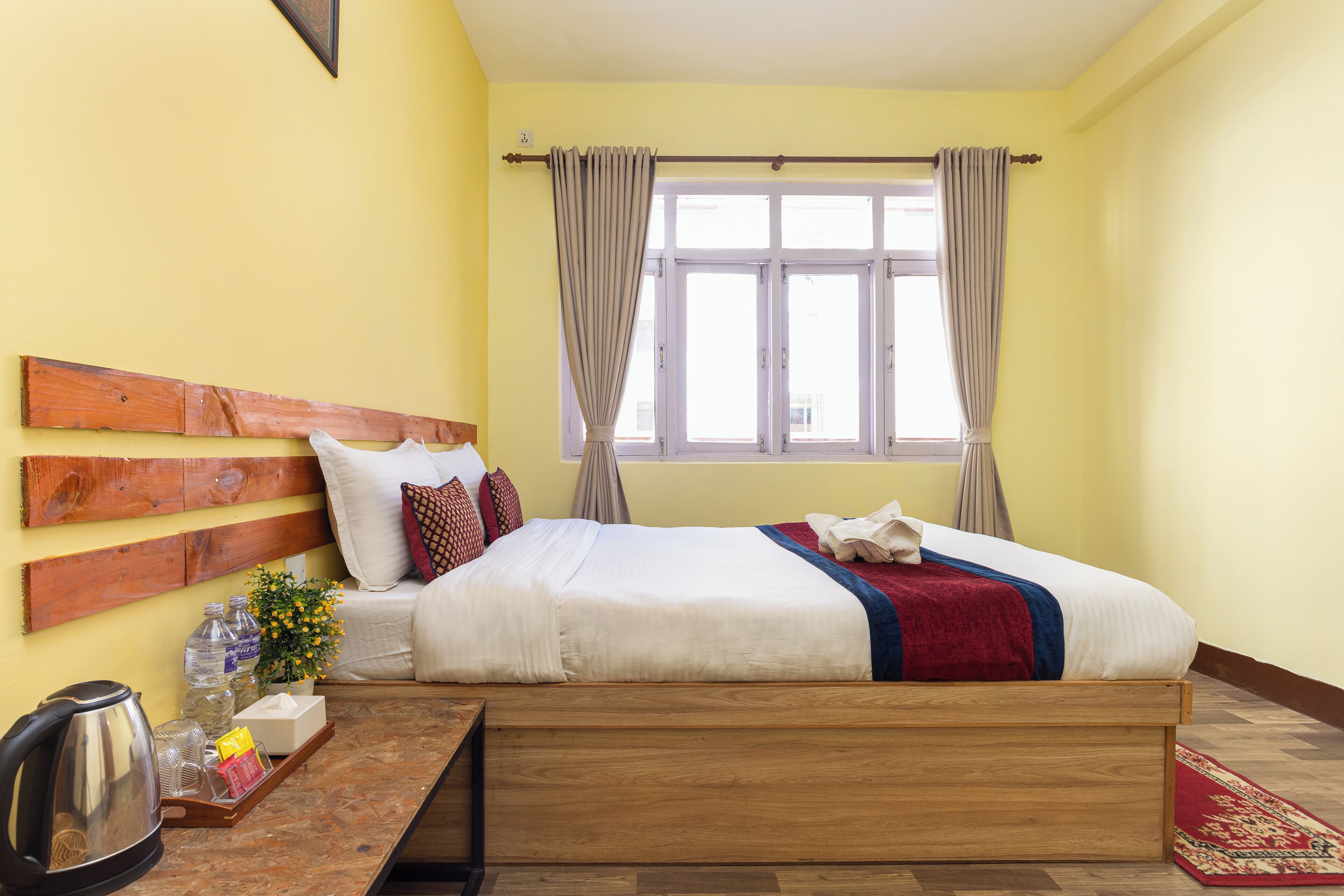 Habitación de hotel ordenada con cabecera de madera, cama con ropa blanca y una manta roja, mesita de noche de madera con una tetera y botellas, y ventana con cortinas.
