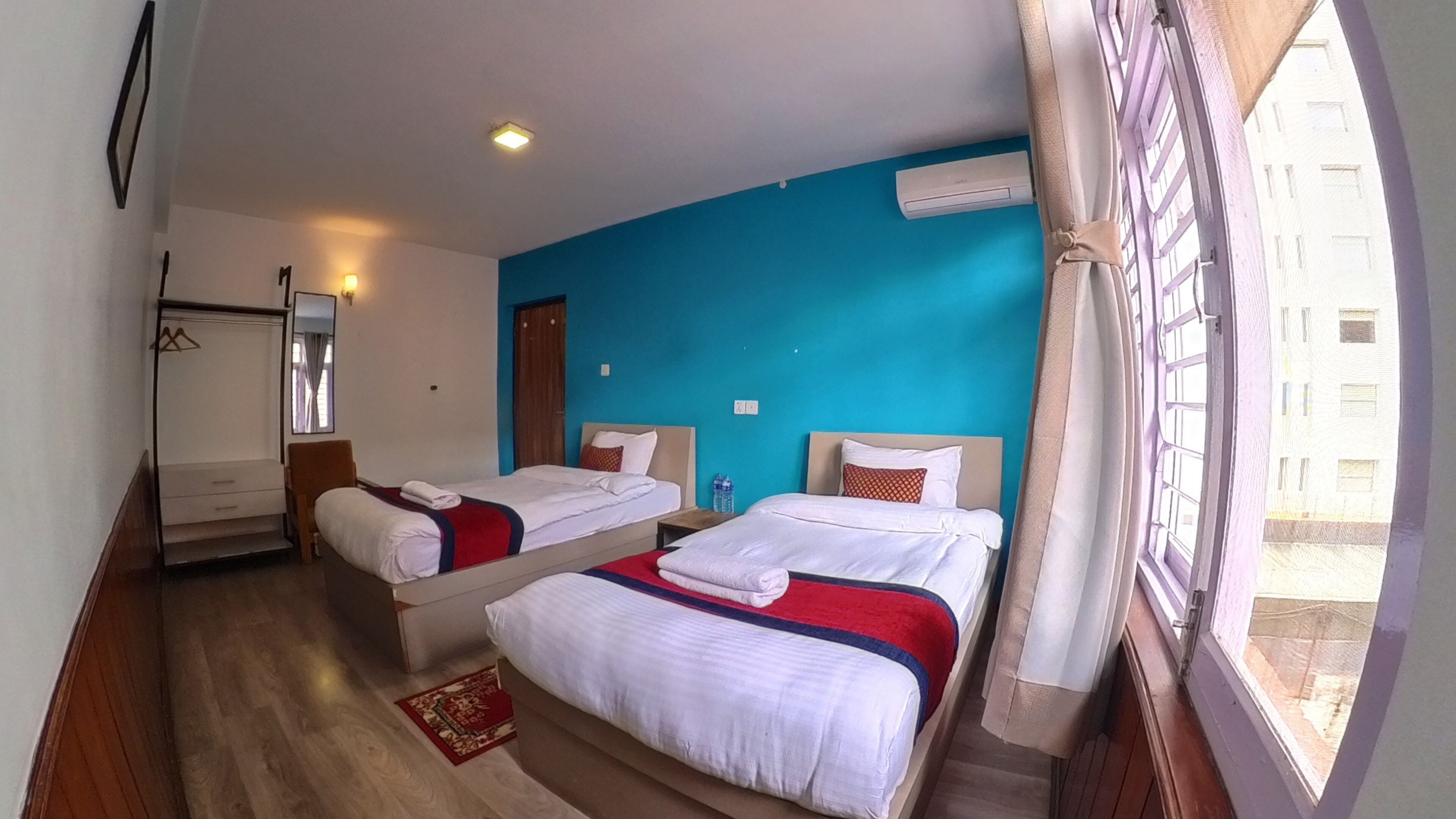 A hotel room with two beds, a wardrobe, a mirror, a window with curtains, and an air conditioning unit on the wall.
