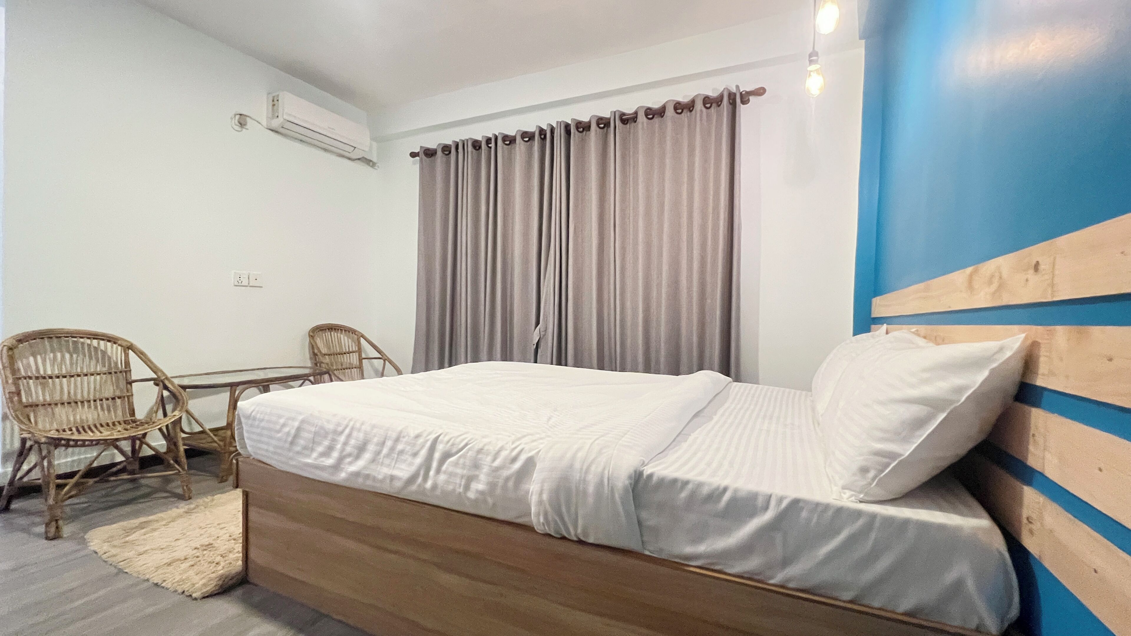 A bedroom with a wooden bed, a small table, two wicker chairs, a grey curtain, and a blue accent wall.