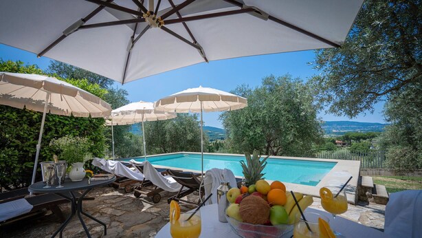 Área da piscina com guarda-sóis, espreguiçadeiras e uma mesa com bebidas e frutas.