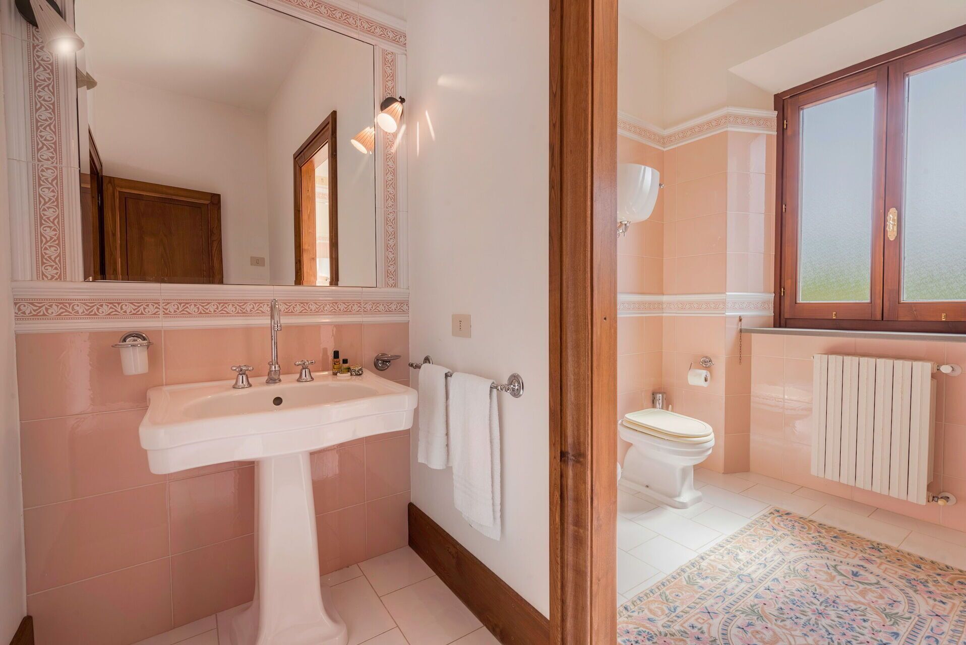 Une salle de bain avec un lavabo, un miroir et des toilettes blancs, des carreaux roses et un tapis à motifs.