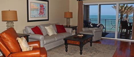 A living room with a sofa, armchair, and coffee table, a ceiling fan, and a view of the ocean through large sliding doors.