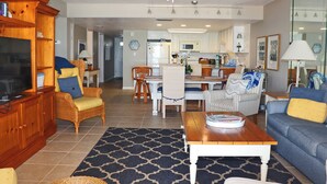 A spacious living room with a wooden entertainment center, a dining area with a table and chairs, and a kitchen in the background. - Second floor three bedroom three bath direct oceanfront (New Smyrna Beach)