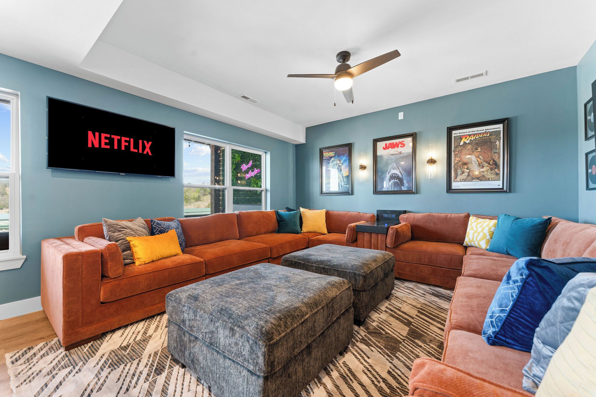 A modern living room with a sectional sofa, ottomans, a flat-screen TV, and framed posters on the wall.