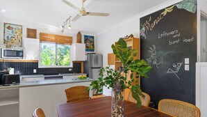 A modern kitchen with a dining area, featuring a chalkboard menu, a ceiling fan, and a large window with a view of greenery. - Enchanted Garden Island Cottage with Bikes and Air Con. (Russell Island)