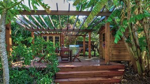 A wooden deck with a grill, surrounded by lush greenery and a wooden structure. - Enchanted Garden Island Cottage with Bikes and Air Con. (Russell Island)