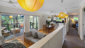 A spacious living area with a sofa, dining table, and chairs, a kitchen with a microwave, and a balcony with a view of greenery. - Enchanted Garden Island Cottage with Bikes and Air Con. (Russell Island)