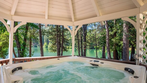A hot tub under a wooden gazebo with a view of a lake and trees. - The Lake House  - Beachfront w/ Hot tub available! (Arnold)