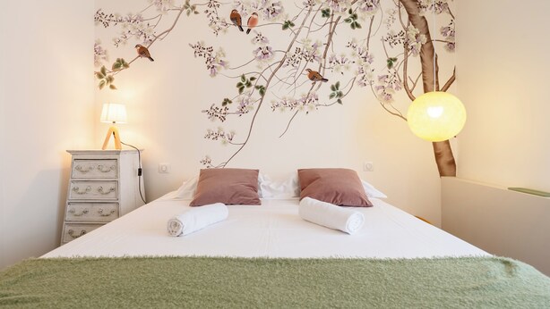 A neatly made bed with two pillows, a green throw blanket, a wall-mounted lamp, and a dresser with drawers.