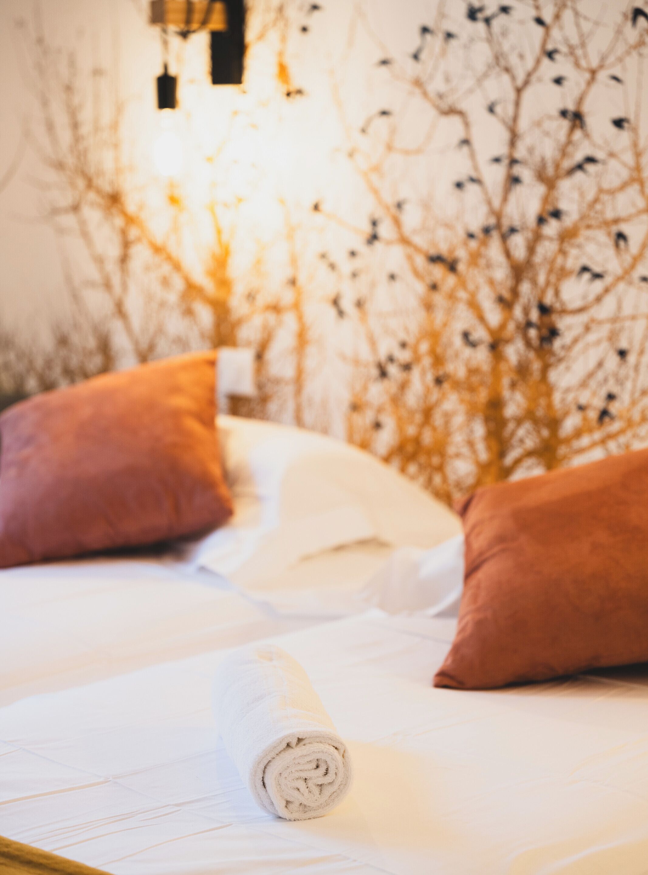 A bed with white linens, two brown pillows, and a rolled white towel.