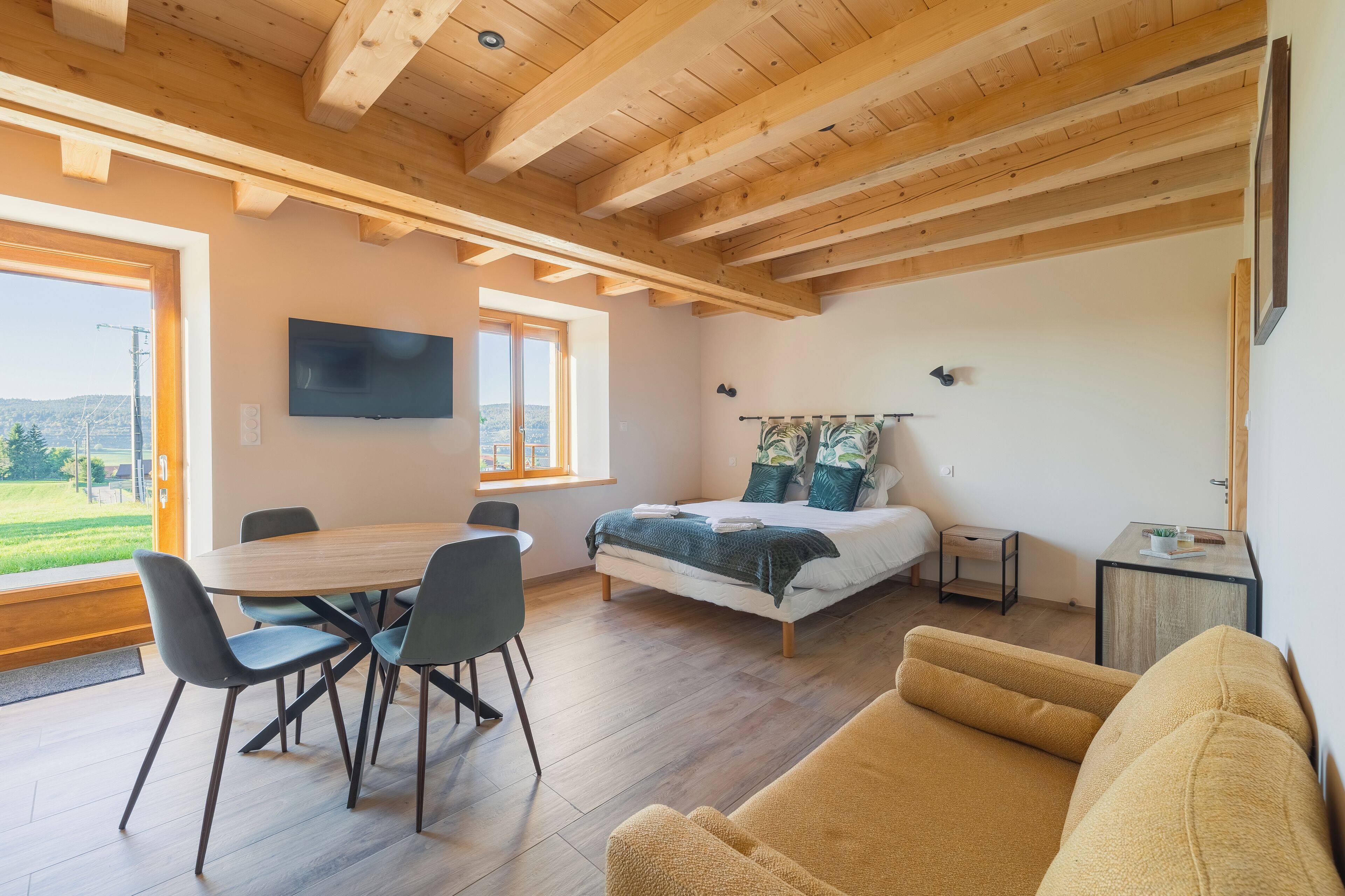 A modern room with a wooden ceiling, a bed with a green blanket, a TV, a dining table with chairs, and a sofa.