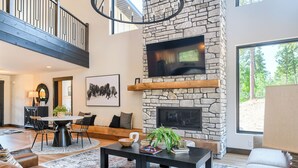 A spacious living room with a stone fireplace, a large TV mounted above it, and a view of the outdoors through a large window. - Modern Spacious Cabin in Powder House w/ Hot Tub, Clubhouse Access to Pools! (Lead)