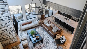 A spacious living area with a stone wall, a large sofa, a coffee table, and a dining area with a long table and bar stools. - Modern Spacious Cabin in Powder House w/ Hot Tub, Clubhouse Access to Pools! (Lead)