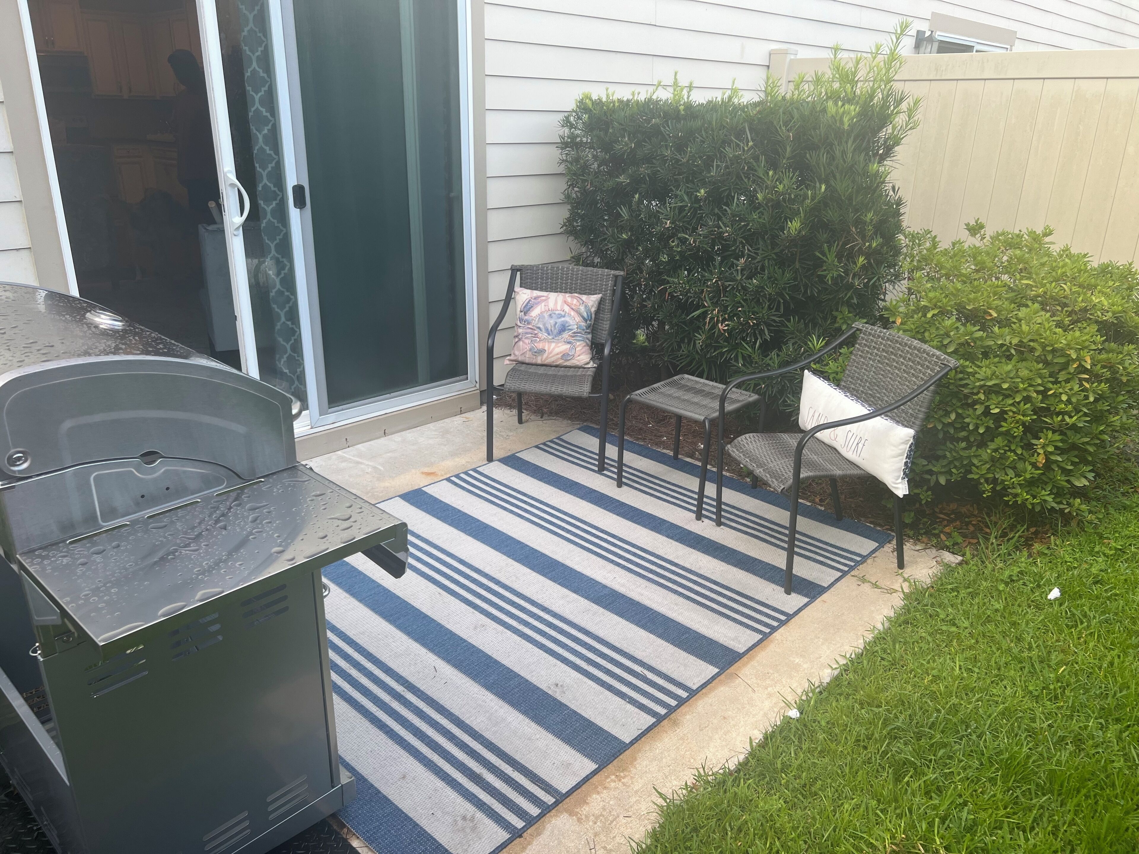 A patio area with a grill, two wicker chairs, a striped rug, and a small bush.