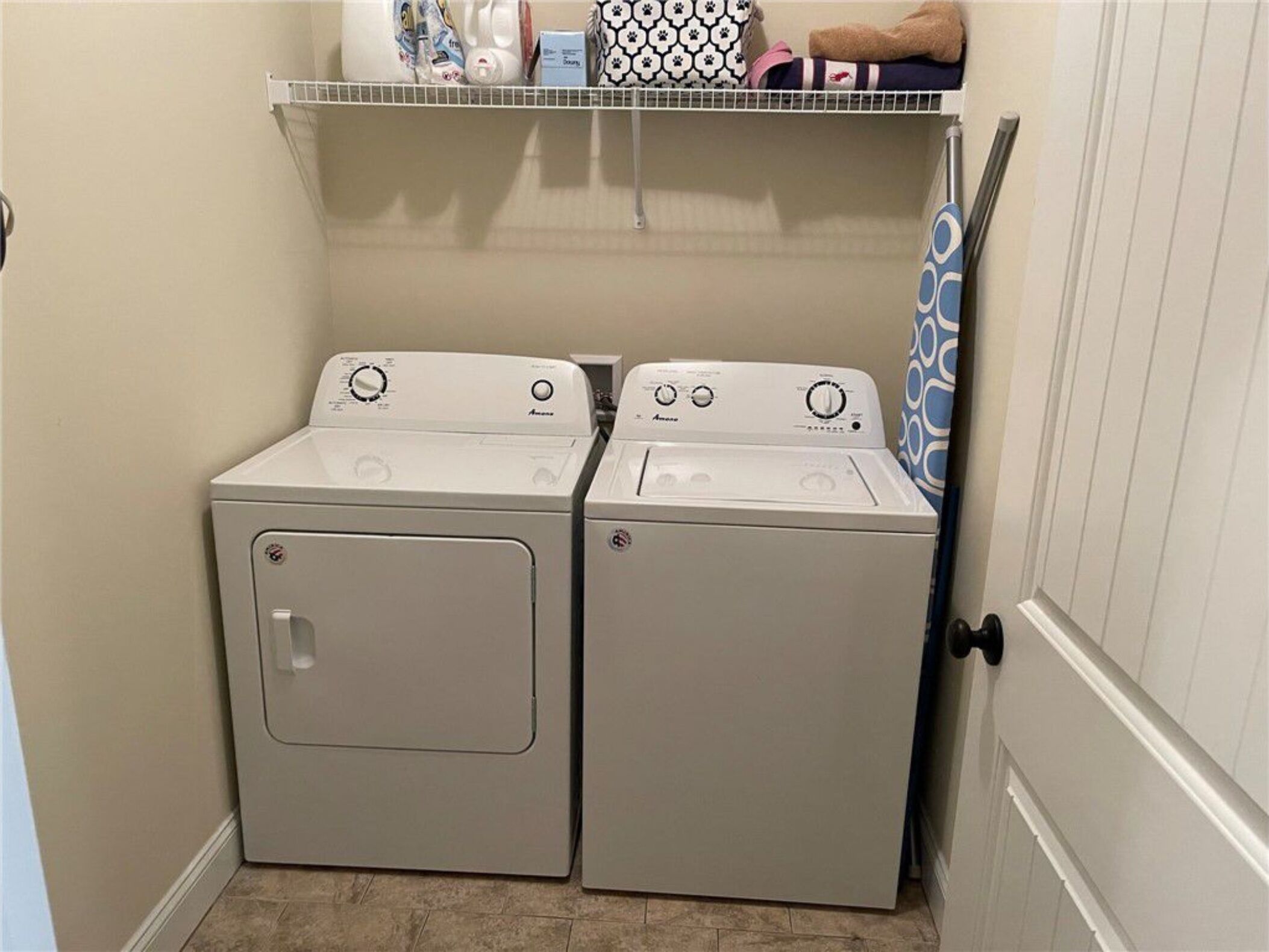 A laundry area with two front-loading washing machines and a shelf above containing various items.