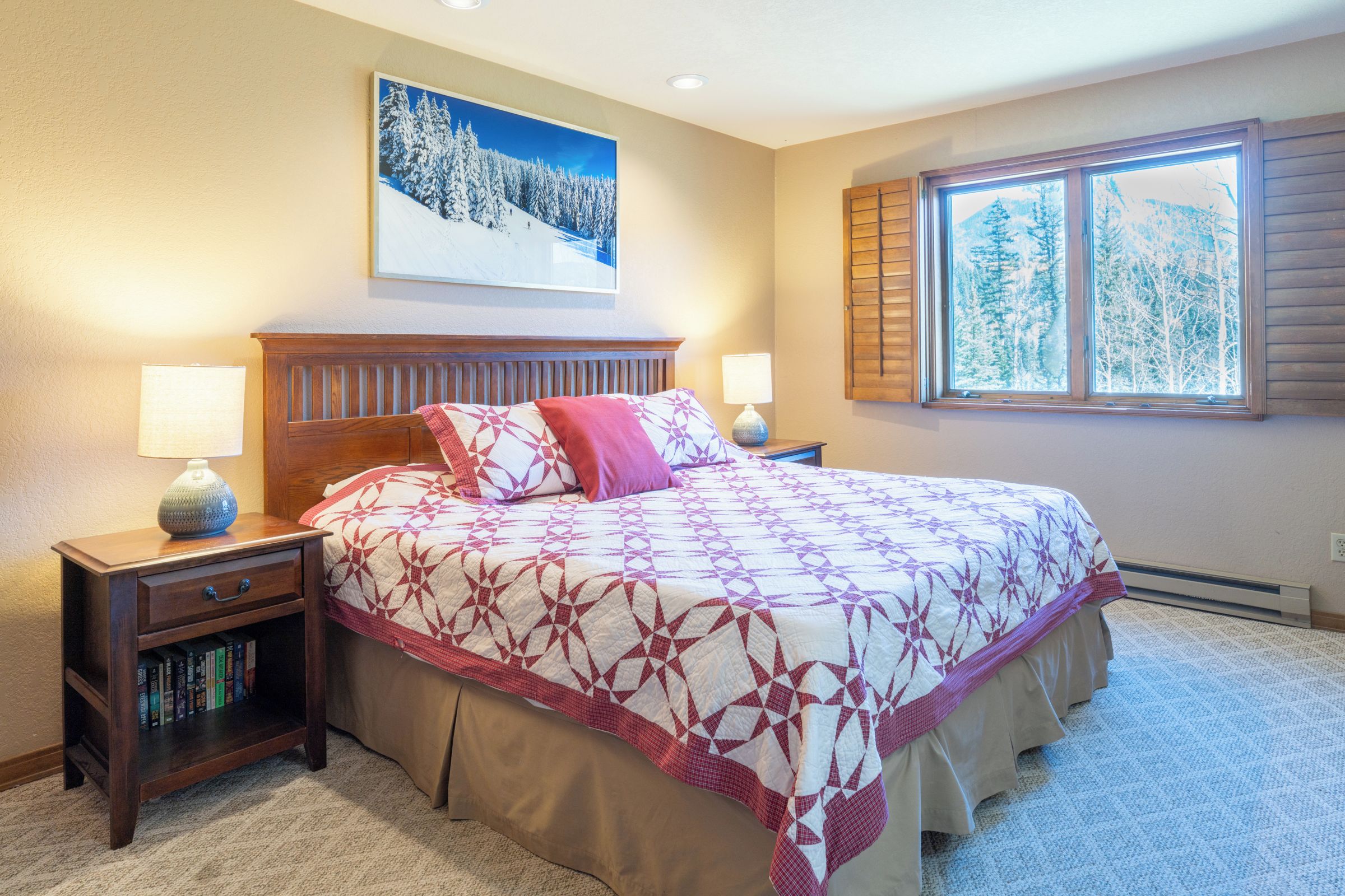 A bedroom with a large bed, two bedside tables with lamps, a bookshelf, a painting of a snowy landscape, and a window with a view of trees.