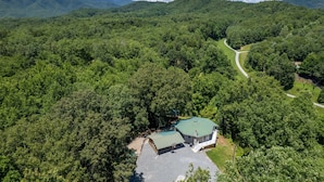An aerial view of a property surrounded by dense forest, featuring a main building with a green roof and a smaller structure, connected by a paved path. - Hike Inn Bryson-Game Room - Fire Pit - Private Location -Wrap Around Deck (Bryson City)