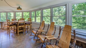 A sunroom with a large table and several wooden chairs, large windows with a view of trees, and a hanging light fixture. - Hike Inn Bryson-Game Room - Fire Pit - Private Location -Wrap Around Deck (Bryson City)