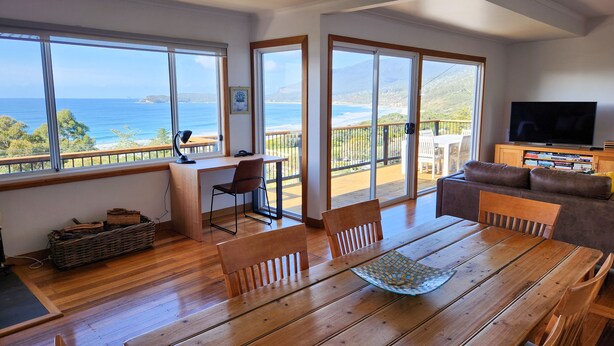 Amplia sala de estar con una mesa de comedor de madera, un sofá y un televisor. Ventanas grandes ofrecen vistas al océano y las colinas.