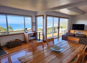 A spacious living area with a wooden dining table, a sofa, and a TV. Large windows offer a view of the ocean and hills. - The Lookout at Pirate's Bay. 3BR, 2BA, ocean views, pool table, arcade machine (Eaglehawk Neck)