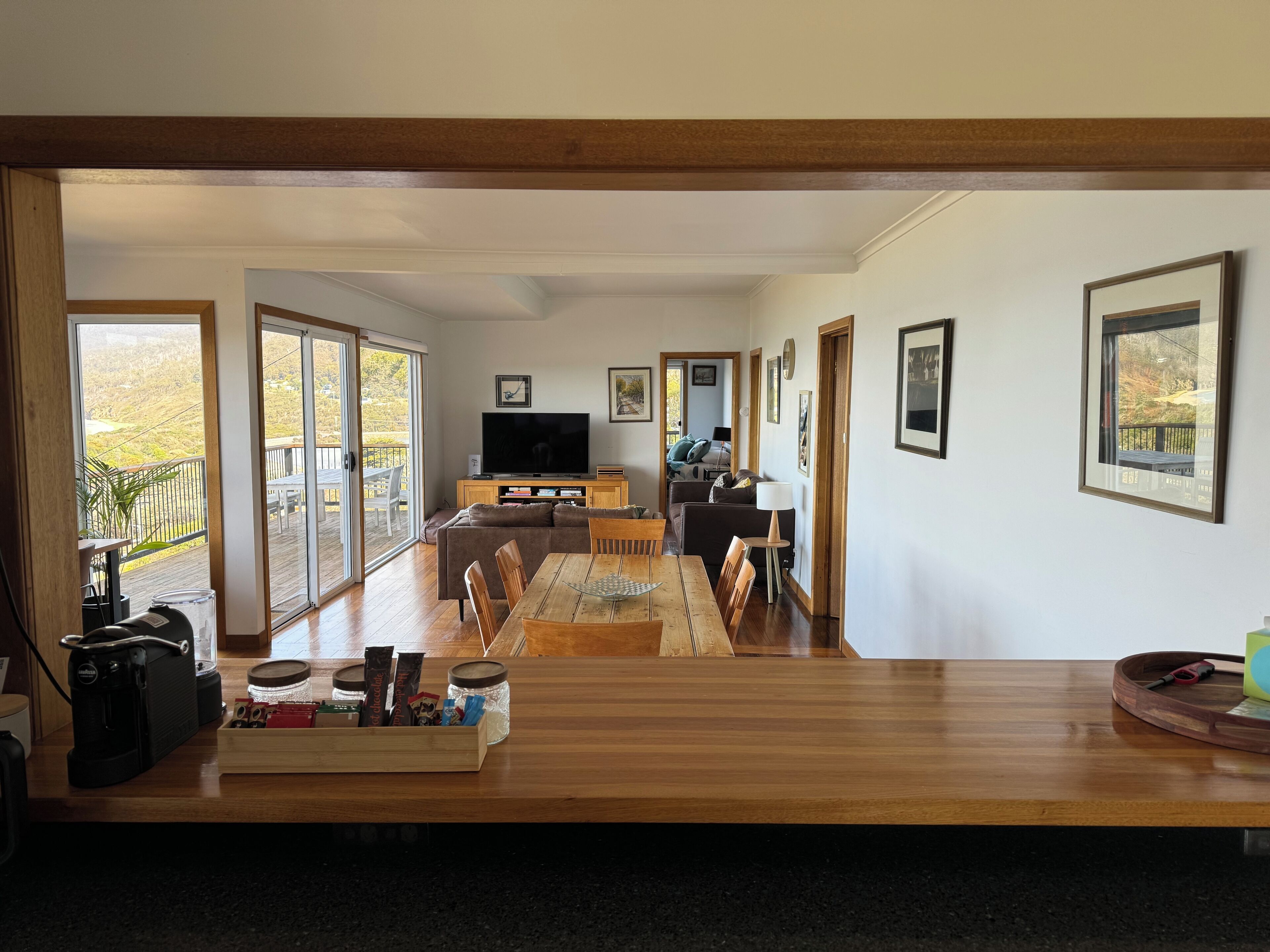 Un amplio salón con una mesa de comedor de madera, un sofá y un televisor. Grandes ventanales ofrecen vistas al exterior.