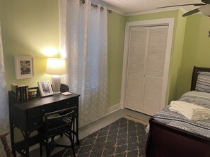Ein Schlafzimmer mit Bett, Schreibtisch mit Stuhl, Lampe, Kleiderschrank und einem gerahmten Bild an der Wand.