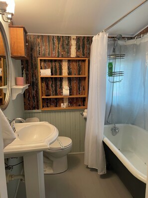 Ein Badezimmer mit weißem Waschbecken, WC und Badewanne. Eine Holzablage hält Toilettenartikel bereit, ein Duschvorhang ist angebracht.