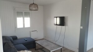 Een woonkamer met een grijze bank, een kleine tafel, een wandtelevisie en een hanglamp.