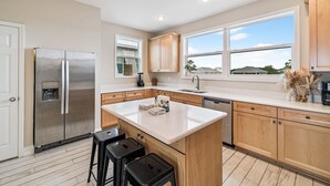A modern kitchen with a stainless steel refrigerator, wooden cabinets, a large window, and a central island with stools. - Beachfront Home w/ Private Boardwalk (Port St. Joe)