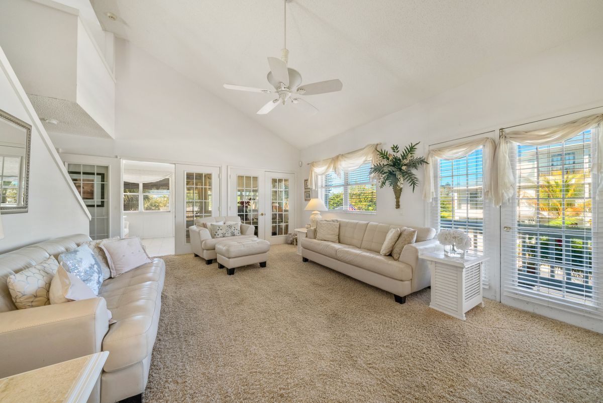 A spacious living room with a beige carpet, white sofas, a ceiling fan, and large windows with curtains.