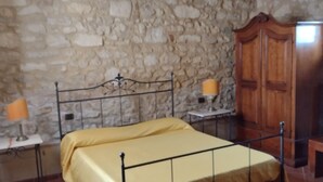 Une chambre avec un mur en pierre, un lit avec un cadre en métal, une armoire en bois et une petite table avec une lampe.