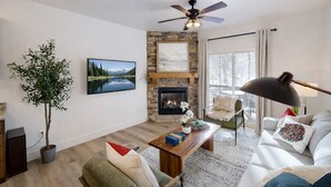A modern living room with a fireplace, a television, a ceiling fan, and a large window. - 5569 Bear Hollow Hideaway! 3 Bedroom Condo by RedAwning (Park City)