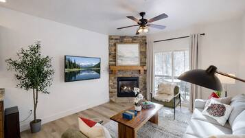 A modern living room with a fireplace, a television, a ceiling fan, and a large window.