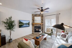 A modern living room with a fireplace, a television, a ceiling fan, and a large window.