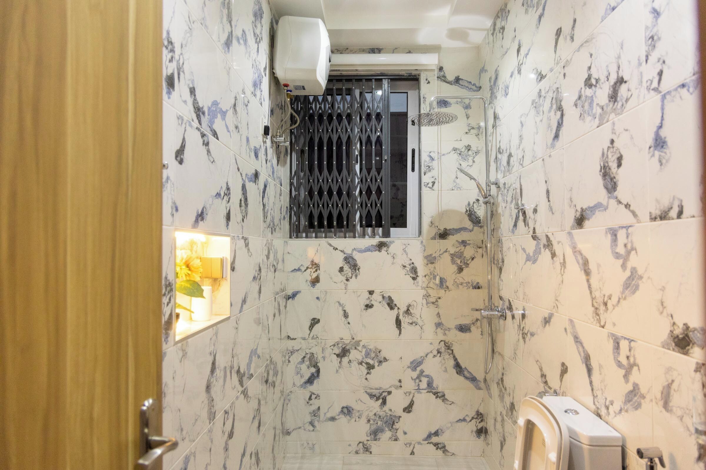 A bathroom with marble tiles, a shower, a toilet, and a window with a metal grille.