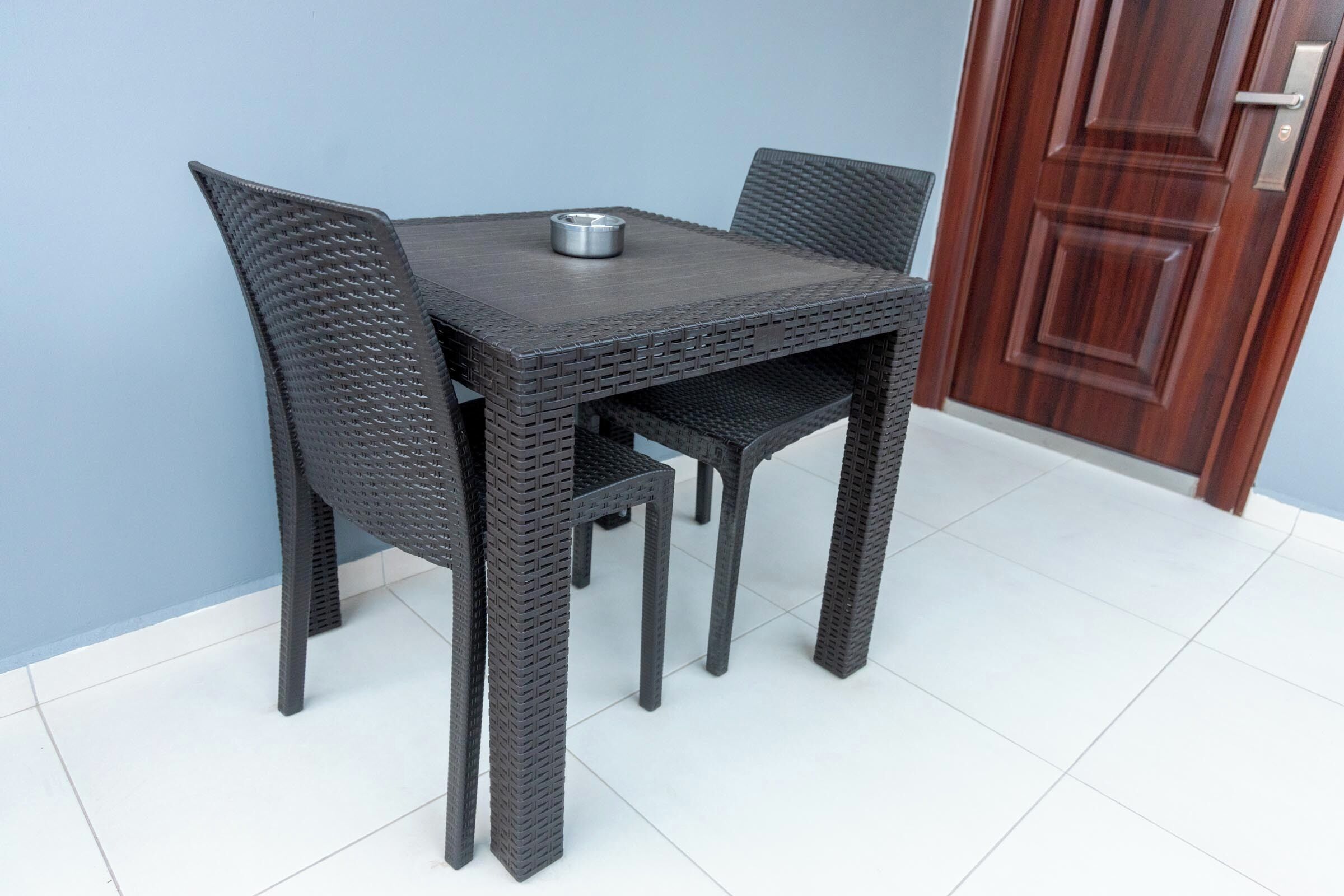 A dark wicker table with two chairs, a metal ashtray, and a wooden door in the background.