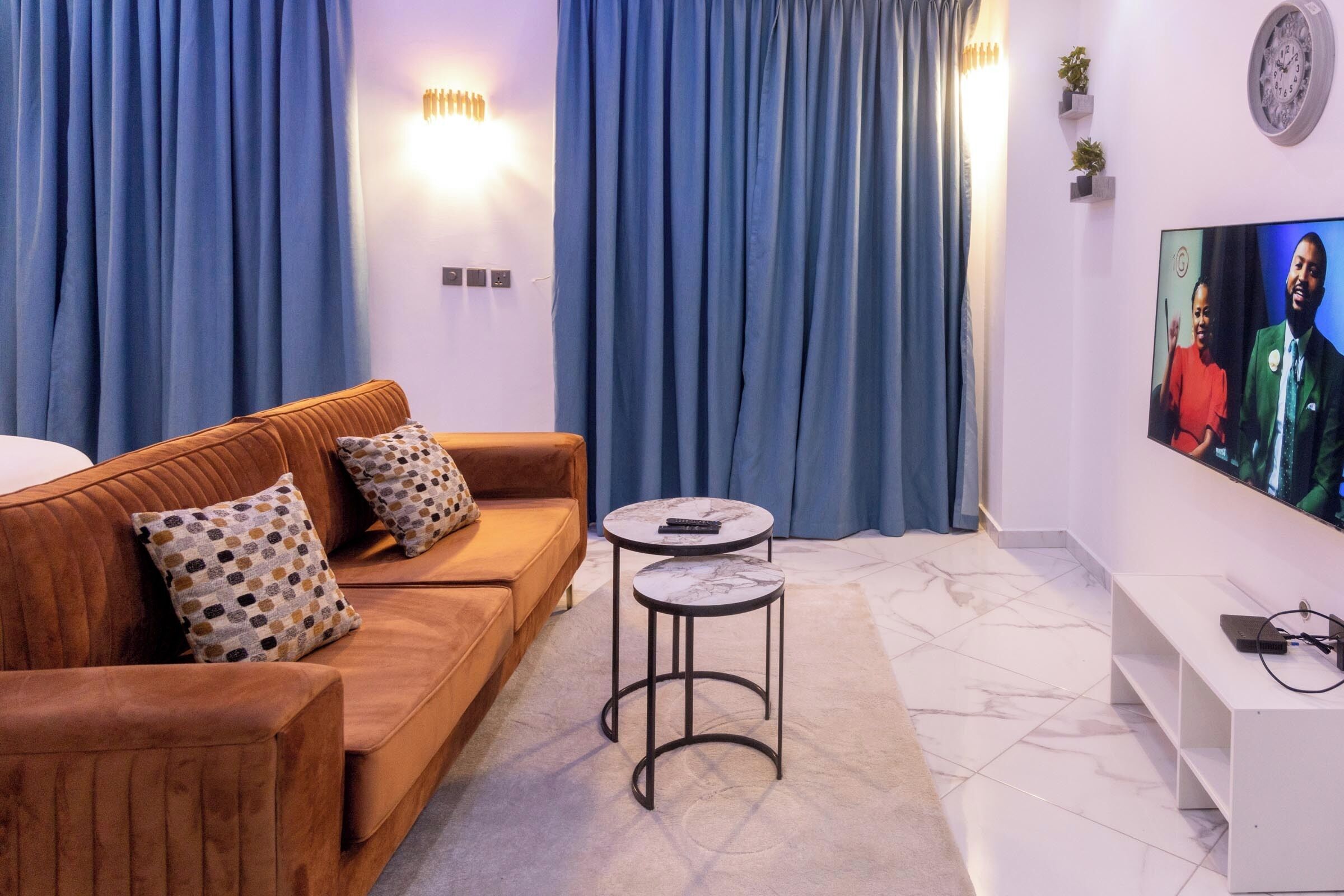 A modern living room with a brown sofa, two side tables, a flat-screen TV, and blue curtains.