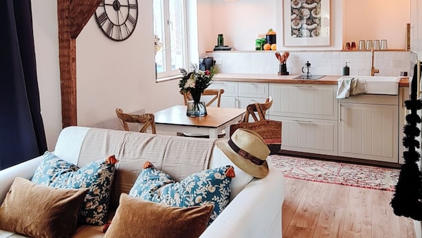 Una sala de estar acogedora con un sofá blanco, suelo de madera y una cocina al fondo.