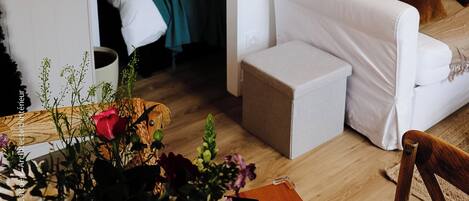 A cozy room with a bed, a white sofa, a small table with a vase of flowers, and a window with a view of the outdoors.