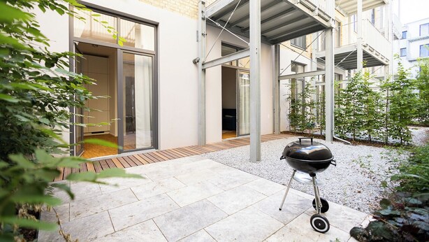 A modern outdoor patio with a barbecue grill, a covered seating area, and a view of a building with large windows.