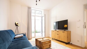 A modern living room with a blue sofa, a wooden coffee table, a TV on a wooden cabinet, and a large window with curtains.
