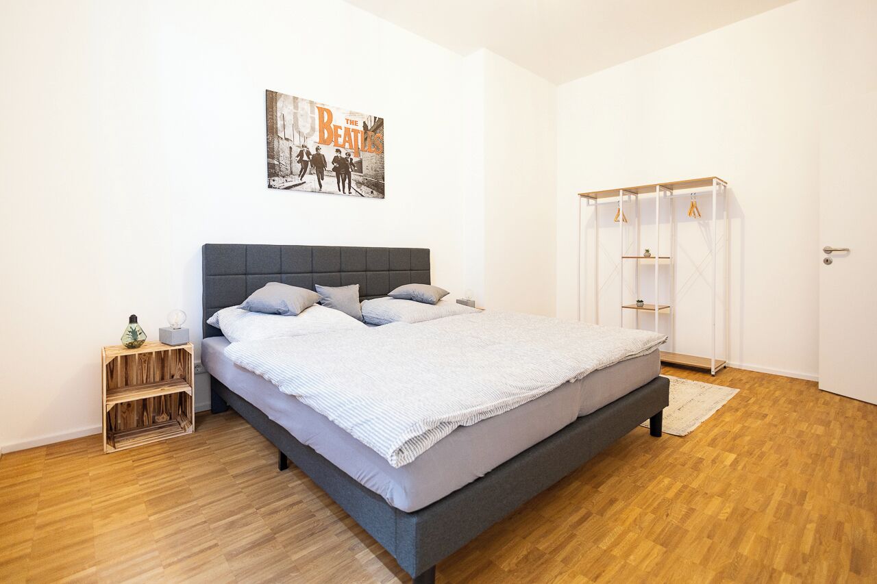 Ein modernes Schlafzimmer mit grauem Bett, einem Holznachttisch, einem an der Wand befestigten Kleiderschrank und einem Beatles-Poster.