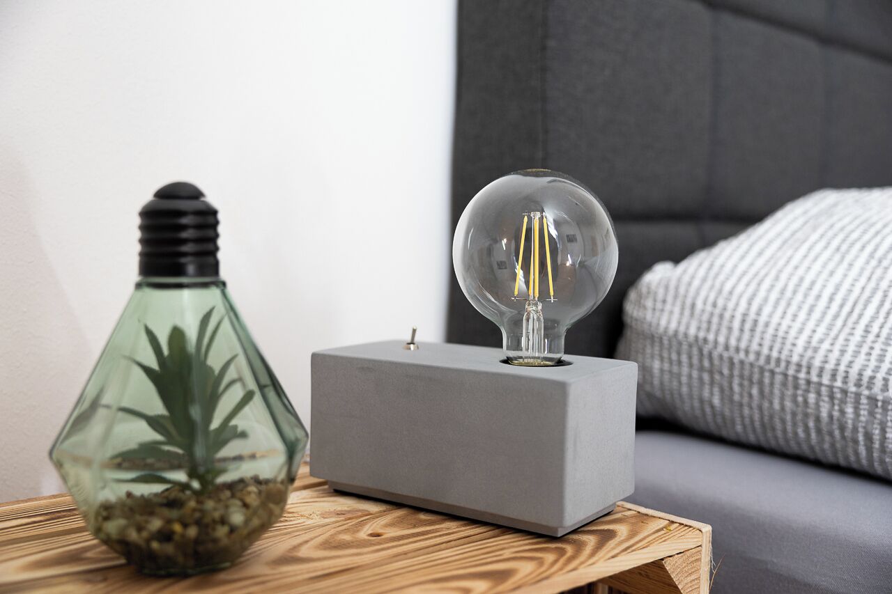 Una camera moderna con una radio grigia squadrata su un tavolino in legno, una lampada in vetro trasparente con una lampadina e una bottiglia di vetro con una pianta.